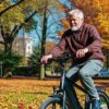 A senior man enjoys a ride on his electric bike, surrounded by vibrant fall foliage and a clear blue sky.